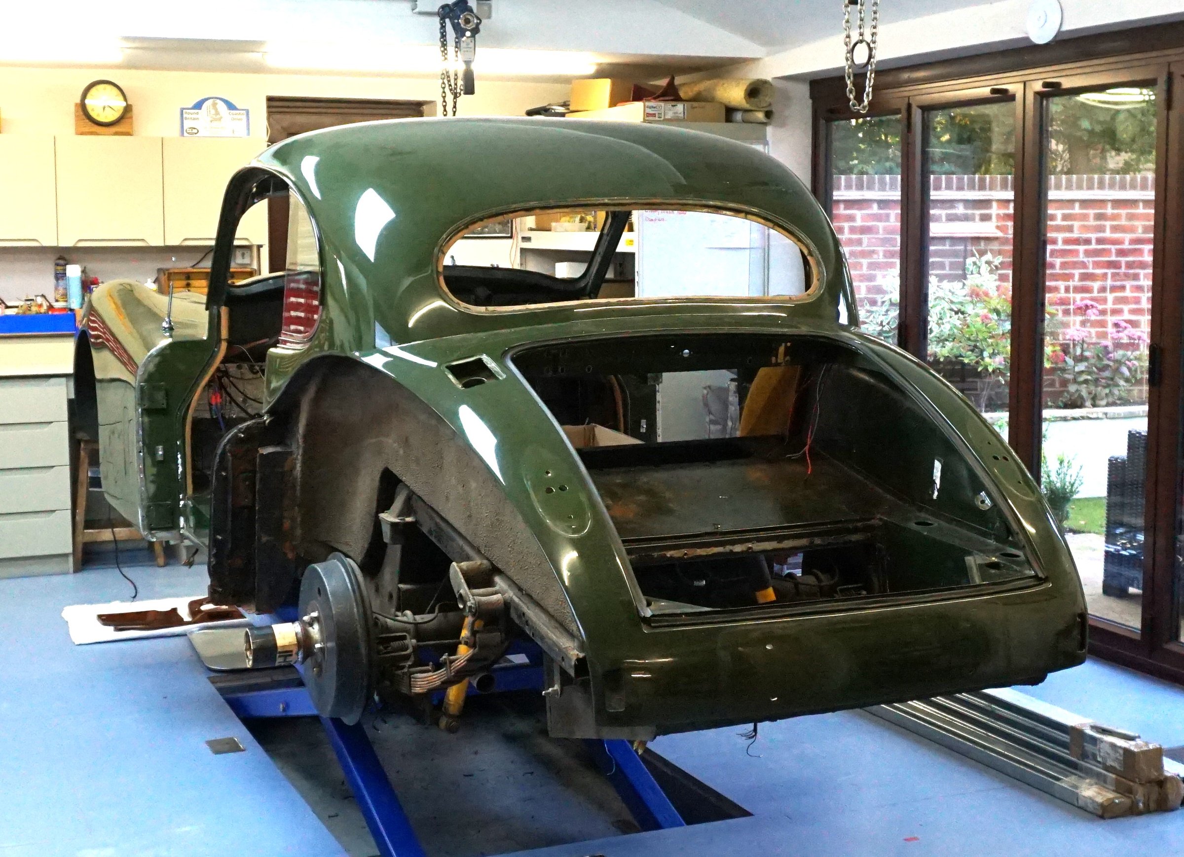 A Jaguar XK140 with its doors and boot lid removed, raised on a four-post lift in a domestic garage, with a chain hoist above it