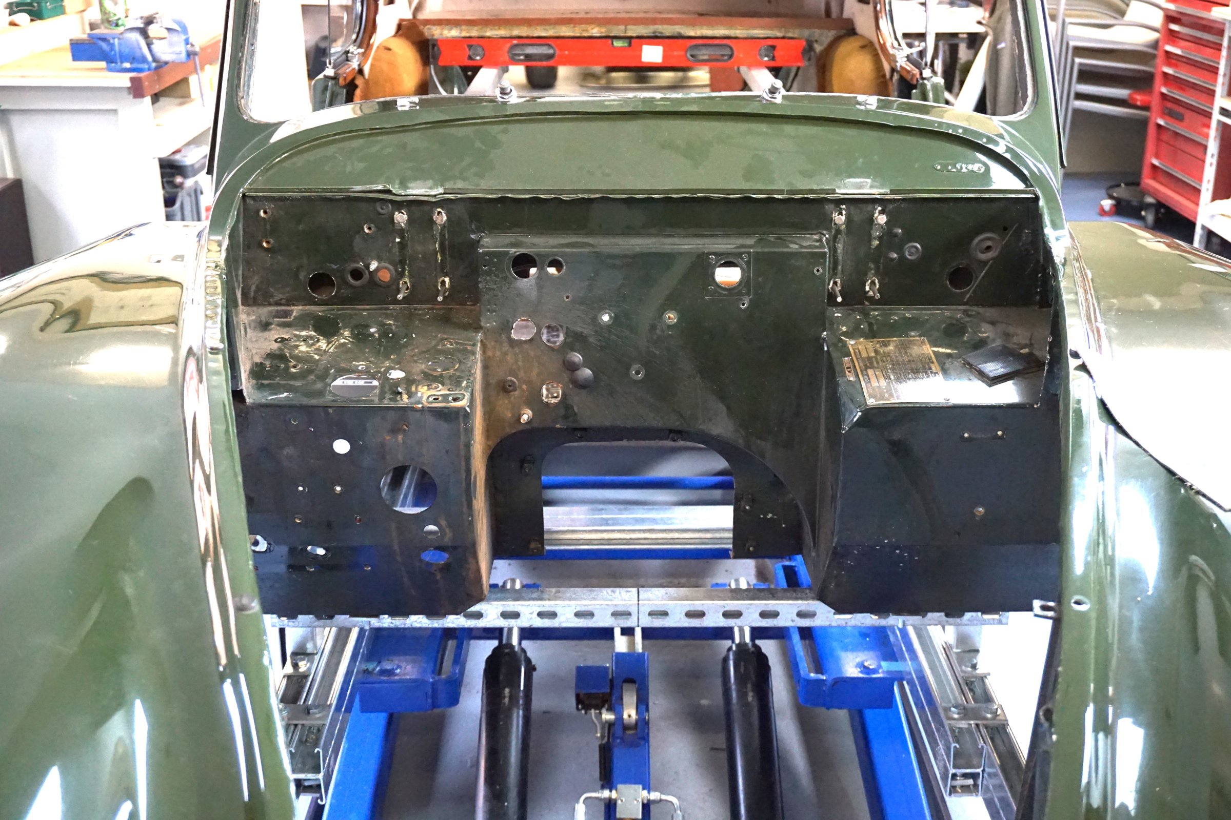 The XK140 body shell lifted off its chassis and carried on galvanised strut across a lift, seen from the front looking at the bare bulkhead